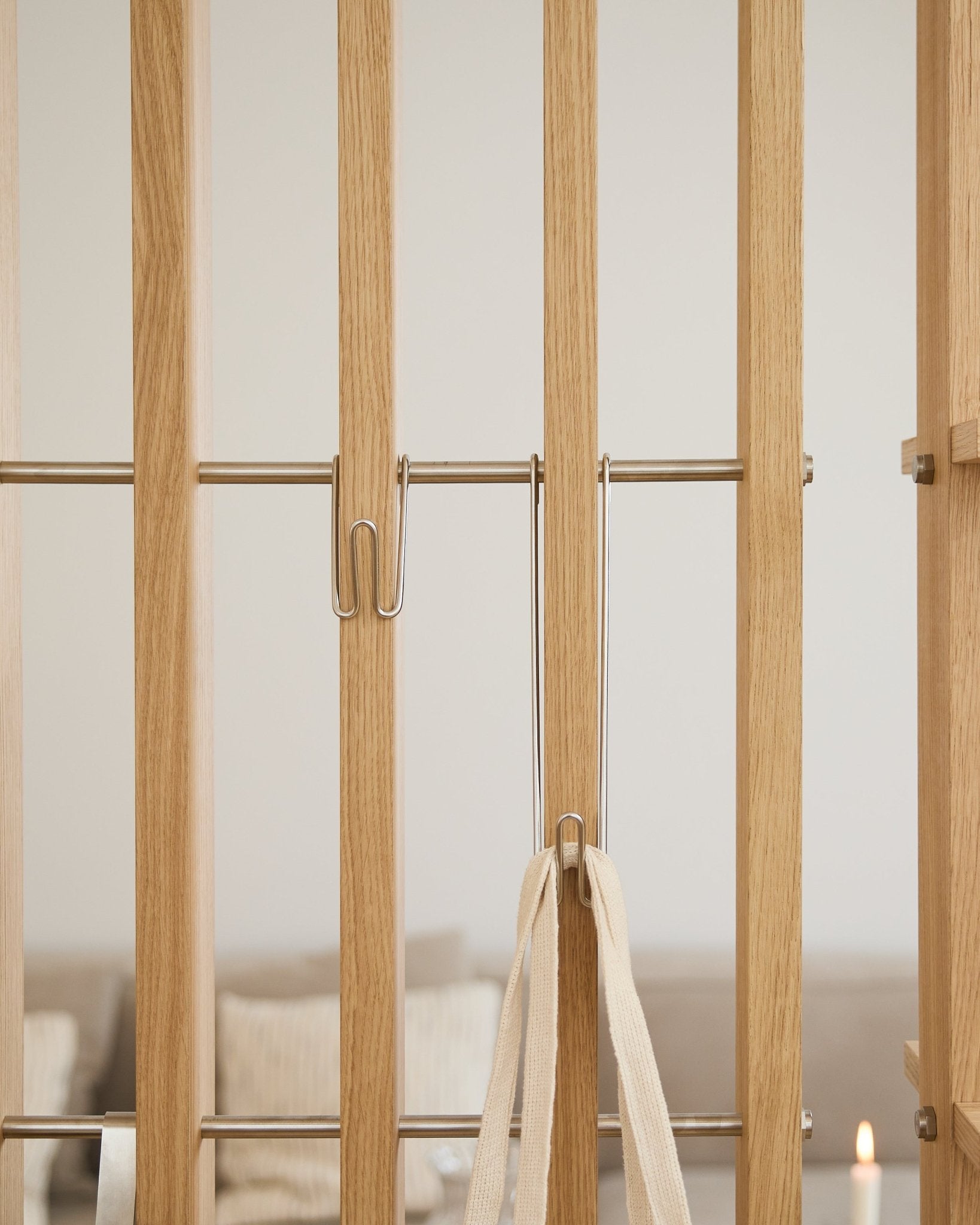 Nærbilde av WoodUpp Alva | Wire Hook BM1 (rustfritt steel, lang, sett med 3) festet til lyse Akustikkpanel spiler med en hvit stropp; sofa og lys er uskarpe i bakgrunnen.
